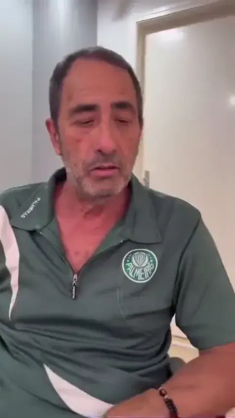 Homem sentado, falando diante da câmera, em uma sala com armário branco ao fundo.