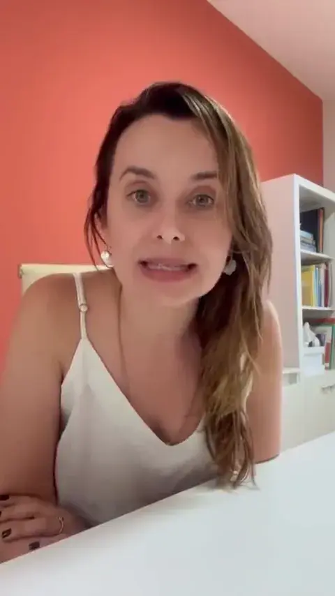 Fonoaudióloga falando diante da câmera, em uma sala com parede rosa e estante ao fundo.